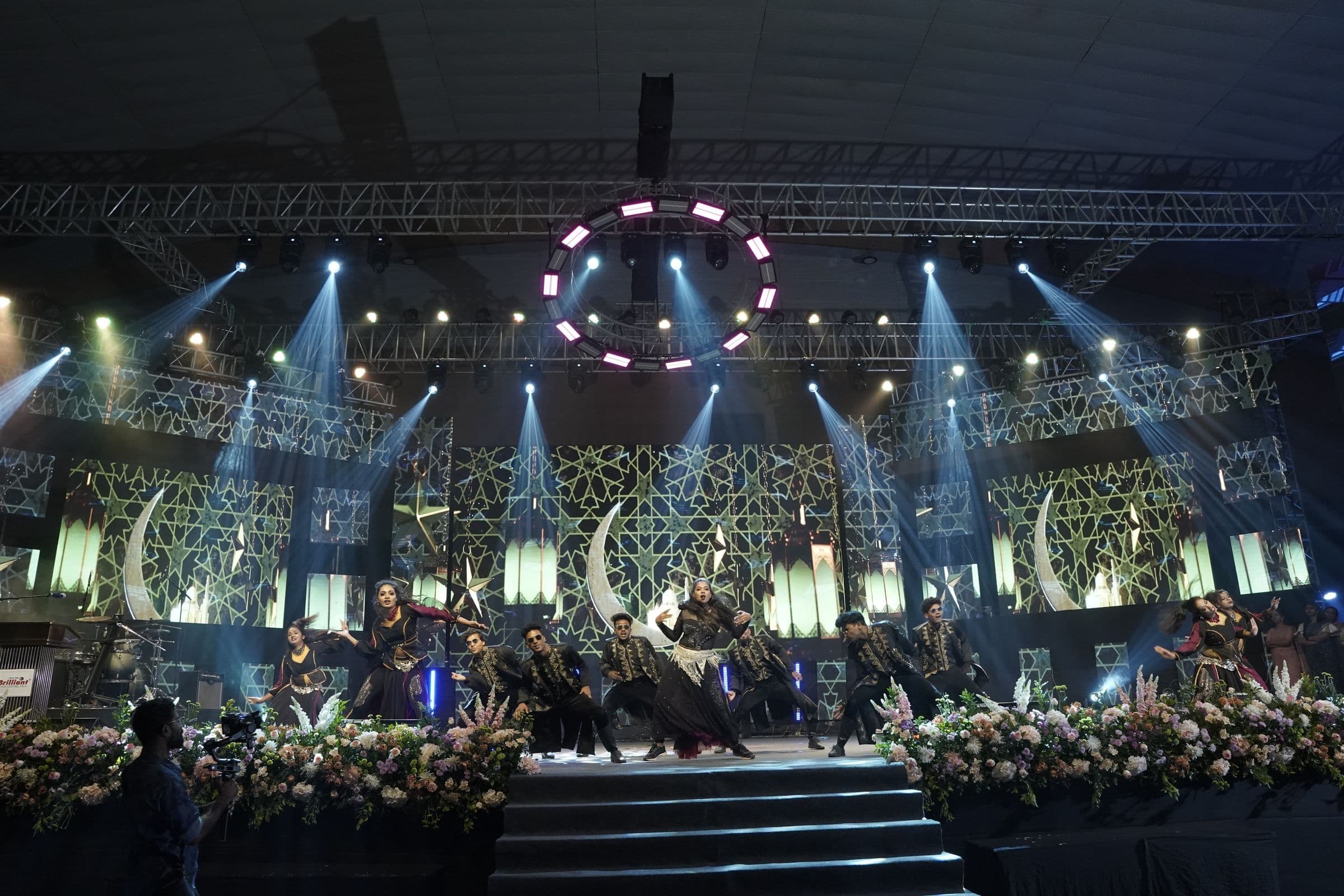 A packed arena stage show lit with moving lights and a ring of LED fixtures