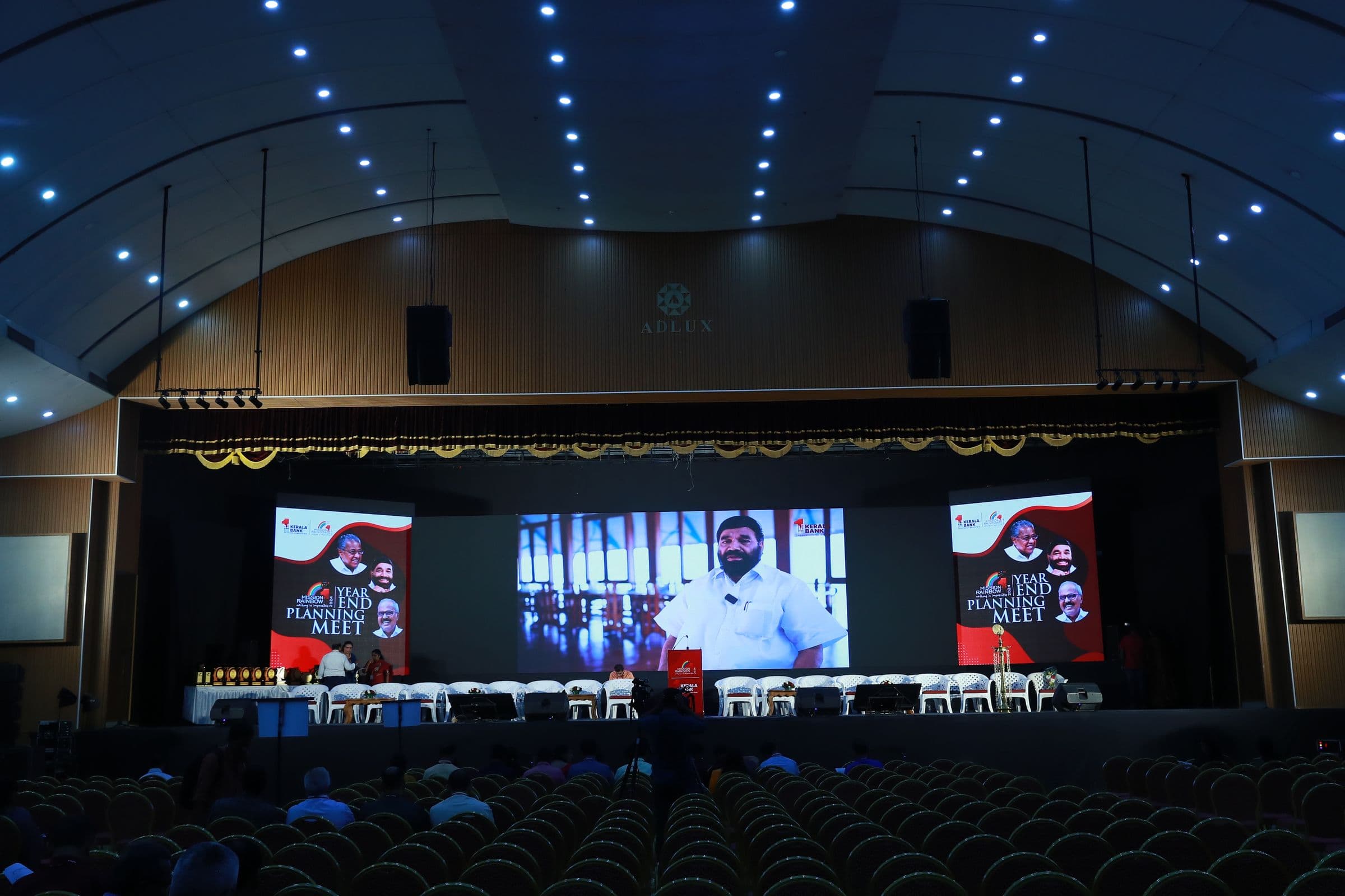 A corporate meet in a full auditorium with speakers relayed on three large screens