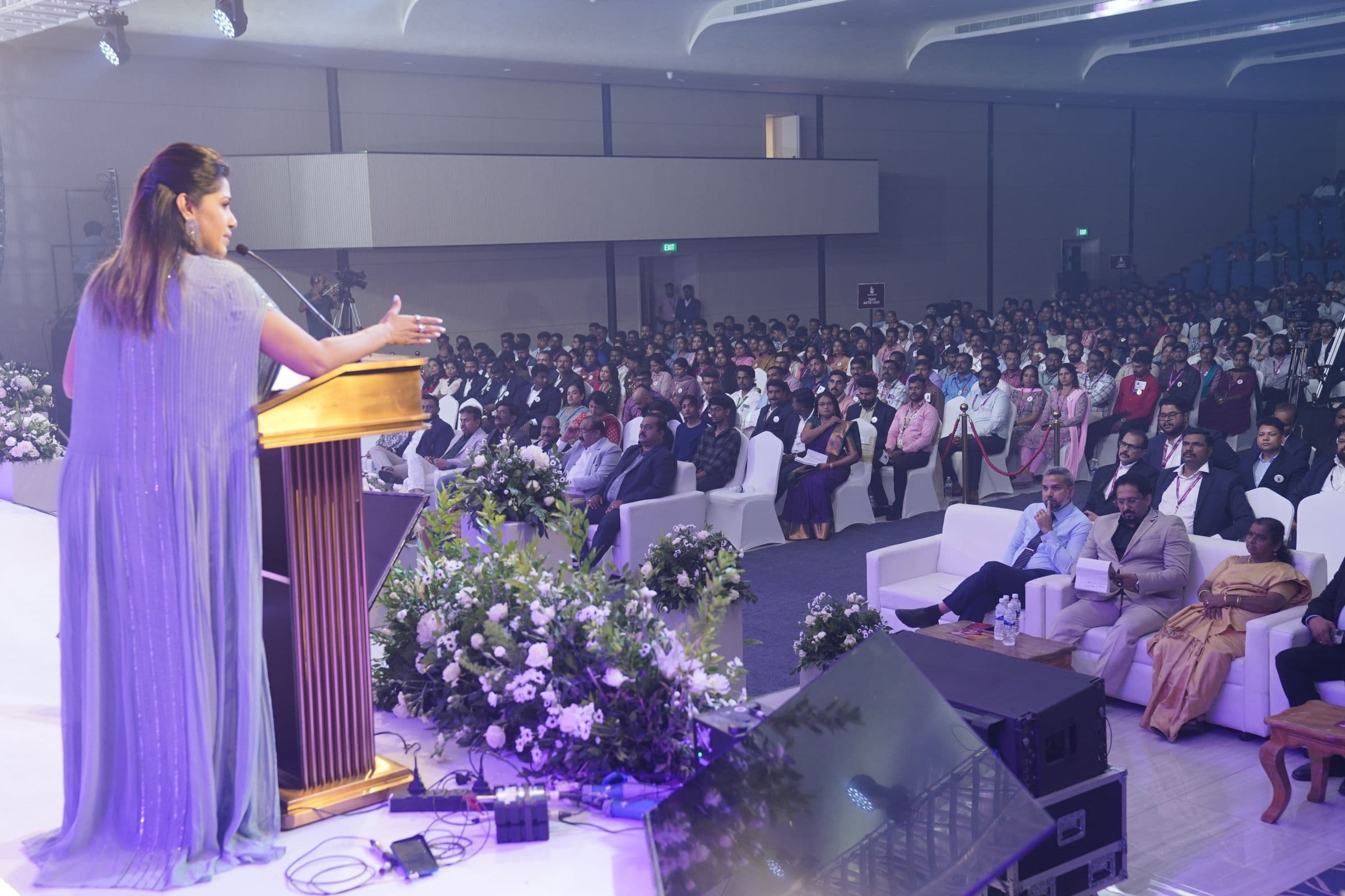 A speaker addresses a seated corporate audience from a floral podium