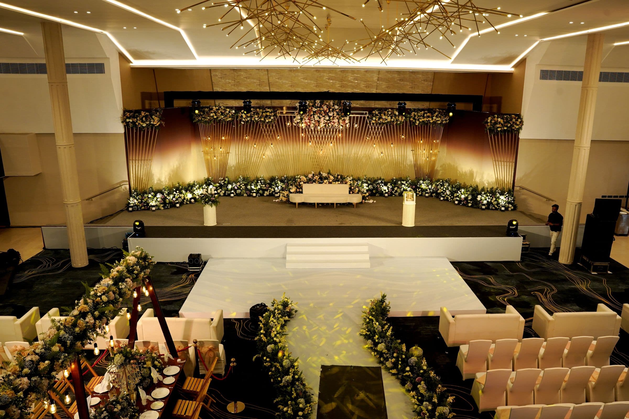 A gold-lit wedding hall with a long floral stage and a white runway