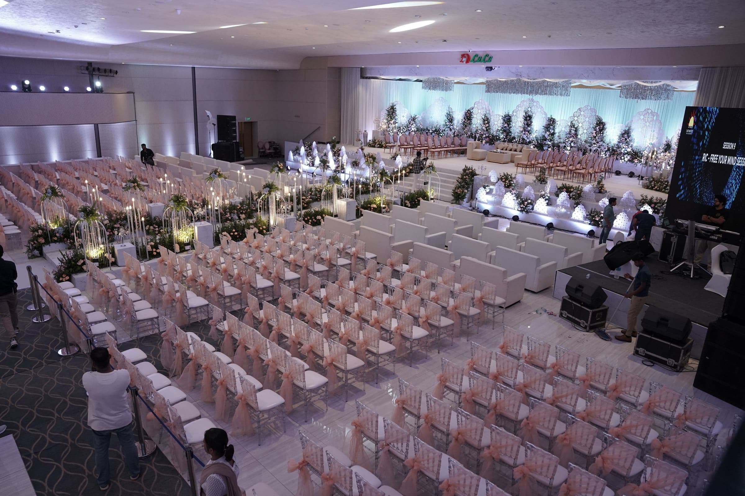 A white-themed wedding hall with rows of dressed seating and floral aisles