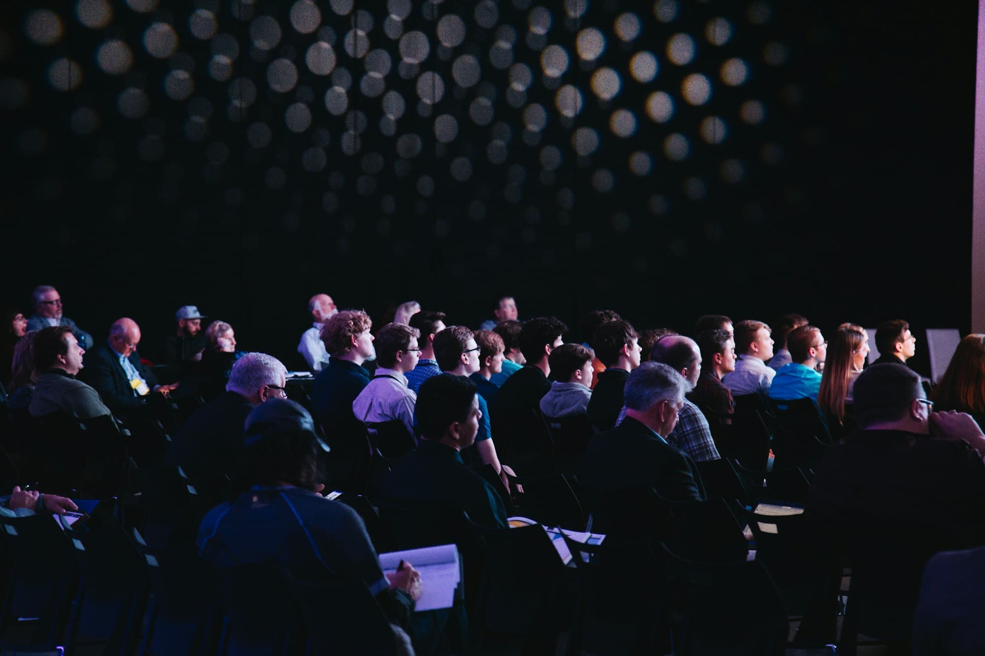 Delegates seated in a darkened conference hall lit by a dotted backdrop